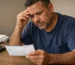 Trabajador puertorriqueño adulto revisando su cheque de pago con expresión de preocupación, sentado en una mesa sencilla en su hogar.