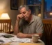 Fotografía editorial realista de una persona adulta puertorriqueña sentada sola en una mesa de comedor durante la noche, revisando facturas y documentos financieros con expresión de agotamiento y reflexión. Sobre la mesa hay calculadora, sobres abiertos y una taza de café. A través de una ventana entra luz tenue de ciudad nocturna. La escena transmite preocupación financiera pero también esperanza y posibilidad de tomar acción.